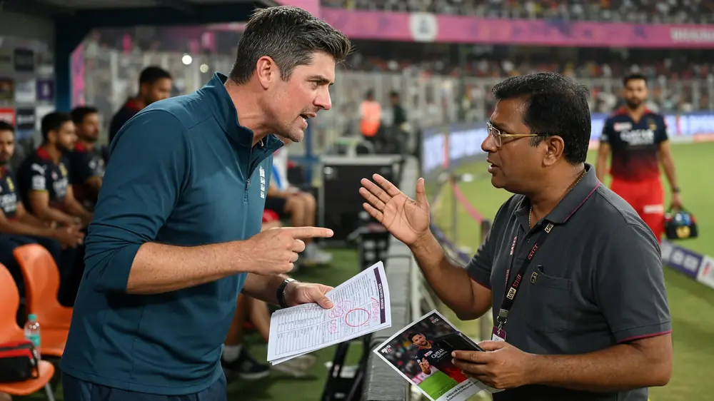A coach in a navy England-style polo points emphatically while debating a bearded staff member in black beside an IPL outfield